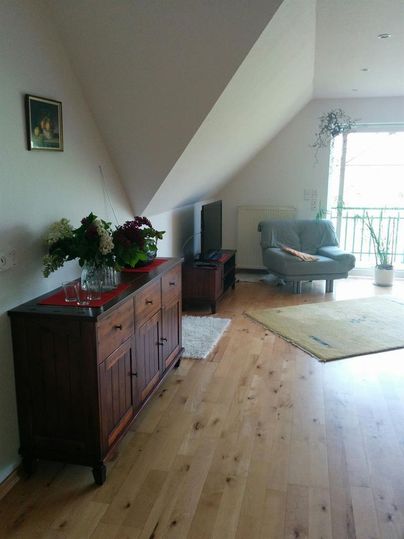 Ein helles Wohnzimmer mit Holzfußboden und schrägen Wänden. Es gibt ein Sideboard, einen Sessel und einen Fernseher, sowie einen Balkon mit Aussicht.