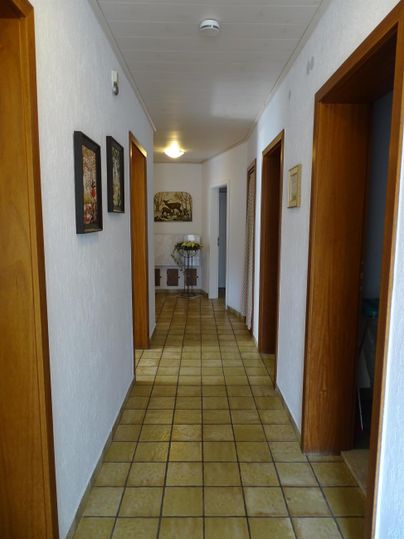 Een lichte gang met houten plafonds en deuren. Aan de muren hangen enkele foto's.