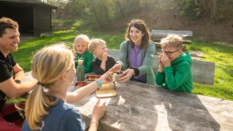 Une famille joyeuse est assise à une table en bois à l'extérieur. Les enfants rient et profitent du temps passé ensemble dans la nature.