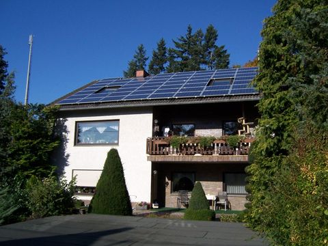 A two-story house with solar panels on the roof. The garden is green and well-maintained, featuring a terrace and an outdoor view.