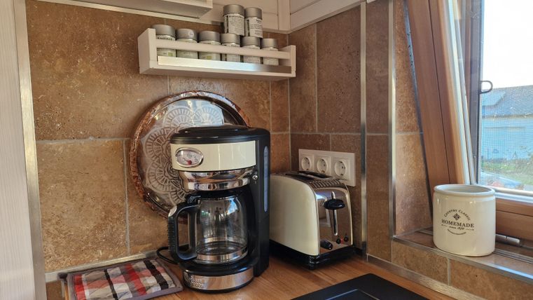 Eine gemütliche Küche mit einer Kaffeemaschine, einem Toaster und Holzarbeitsflächen. Im Hintergrund sind Regale mit Dosen und eine Fensterbank zu sehen.