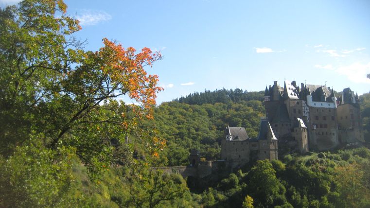 Eine malerische Burg liegt in einer üppig grünen Landschaft. Im Vordergrund sind Bäume mit bunten Blättern zu sehen.