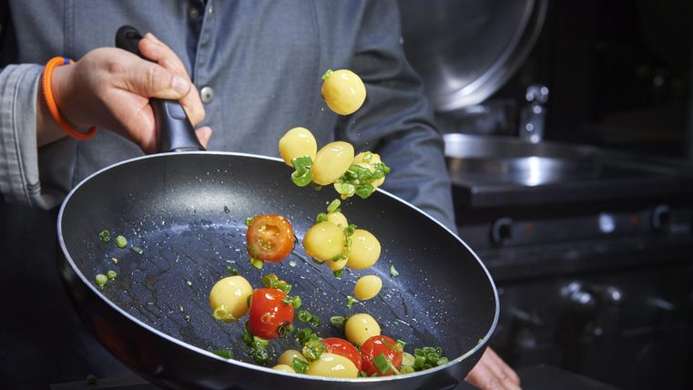 Eine Person brät bunte Gemüse mit einer Pfanne. Verschiedene Zutaten, wie kleine Kartoffeln und Tomaten, springen in die Luft.