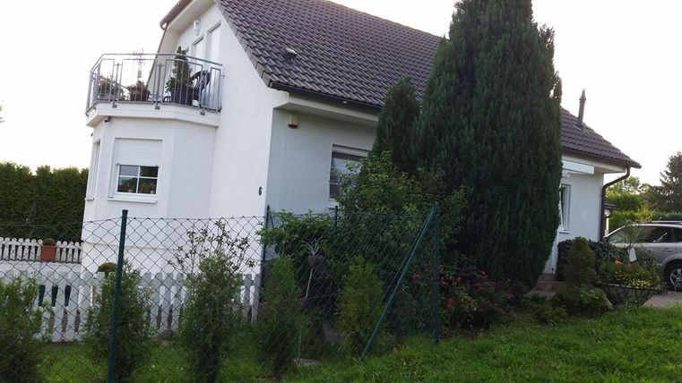 Une maison moderne avec un balcon et un jardin soigné. Entourée d'arbres hauts et d'une clôture.