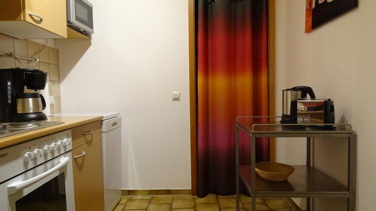 Eine kleine Küche mit Holzschränken, einem Herd und einer Kaffeemaschine. Der Raum hat einen bunten Vorhang und einen Glastisch mit einer Schüssel.