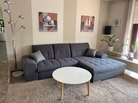Ein modernes Wohnzimmer mit einem grauen Sofa und einem runden Tisch. An den Wänden hängen Kunstwerke, und es gibt einige Pflanzen im Raum.
