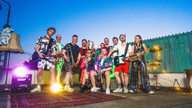 Eine bunte Gruppe von Musikern steht auf einer Bühne unter freiem Himmel. Sie tragen auffällige Kleidung und spielen verschiedene Instrumente bei Sonnenuntergang.