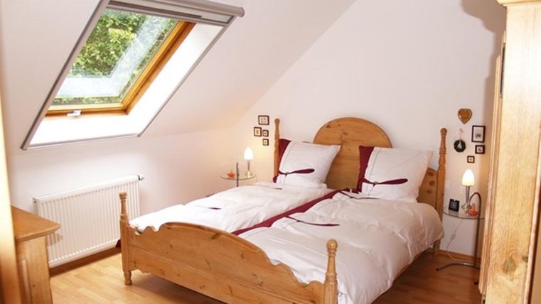 Ein gemütliches Schlafzimmer mit einem Holzbett und zwei weißen Decken. Das Zimmer hat ein Satteldachfenster, das Tageslicht hereinlässt.