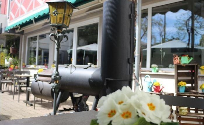 Ein gemütlicher Außenbereich mit Tischen und Stühlen. Im Vordergrund eine gelbe Tasse mit weißen Blumen und im Hintergrund ein großer Grill.