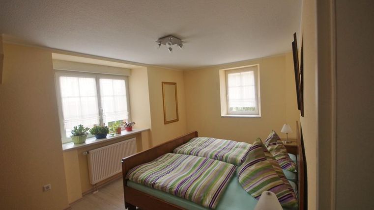 Ein gemütliches Schlafzimmer mit zwei Betten und bunten Bettbezügen. Große Fenster lassen viel Licht herein und Pflanzen sorgen für eine angenehme Atmosphäre.