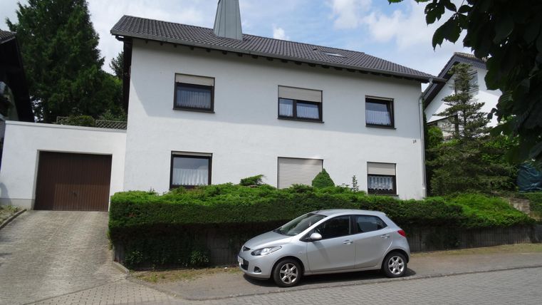 Ein modernes, weißes Einfamilienhaus mit mehreren Fenstern und einem kleinen Vorgarten. Ein silbernes Auto parkt vor dem Haus auf der Straße.