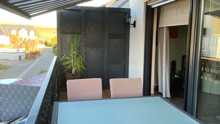 Ein schöner Balkon mit einem Glas-Tisch und vier Stühlen aus Rattan. Im Hintergrund sind Pflanzen und eine verschattete Überdachung zu sehen.
