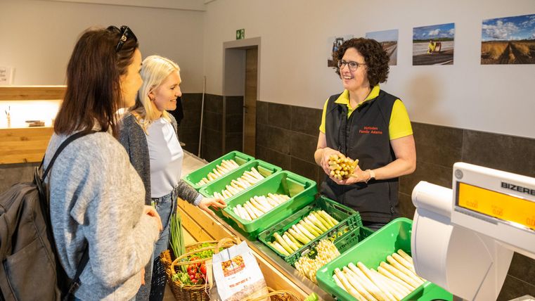 Eine Verkäuferin steht an einem Marktstand und bietet frisches Gemüse an. Zwei Kundinnen sehen sich die angebotenen Produkte an.