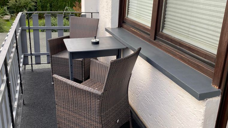 Ein Balkon mit zwei gemütlichen Rattanstühlen und einem kleinen Tisch. Die Umgebung ist grün und gut gepflegt.