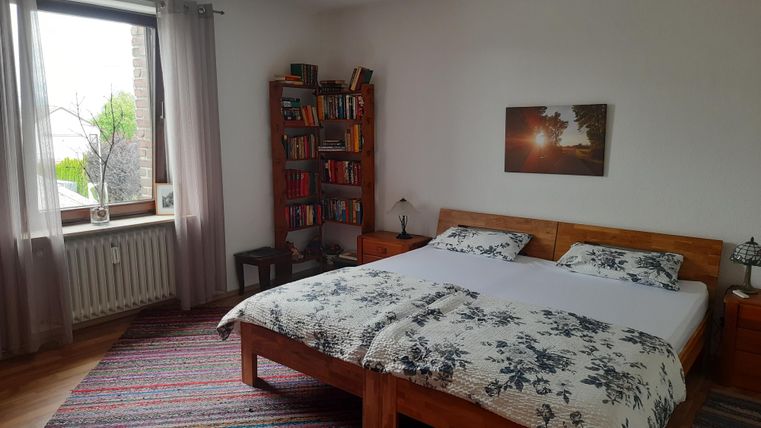 Ein gemütliches Schlafzimmer mit einem großen Bett und buntem Teppich. Im Hintergrund steht ein Bücherregal neben einem Fenster, das Tageslicht hereinlässt.