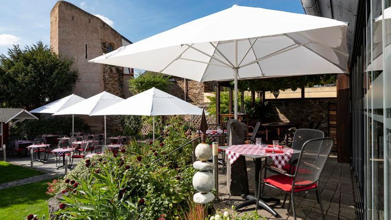 Ein gemütlicher Außenbereich eines Restaurants mit weißen Sonnenschirmen. Der Garten ist grün und einladend, ideal für ein entspanntes Essen im Freien.