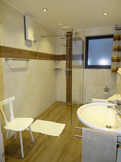 Een moderne badkamer met een douche, een wastafel en een stoel. De wanden zijn licht met houten accenten en er is een raam dat natuurlijk licht binnenlaat.