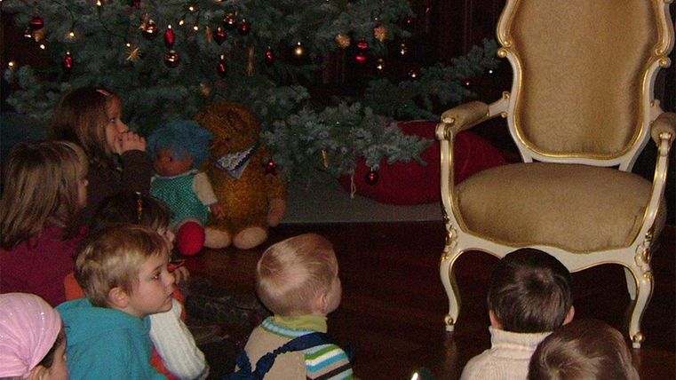 Een groep kinderen zit voor een feestelijk versierde kerstboom en kijkt naar een lege, elegante stoel. Op de achtergrond staat een teddybeer en er heerst een gezellige sfeer.