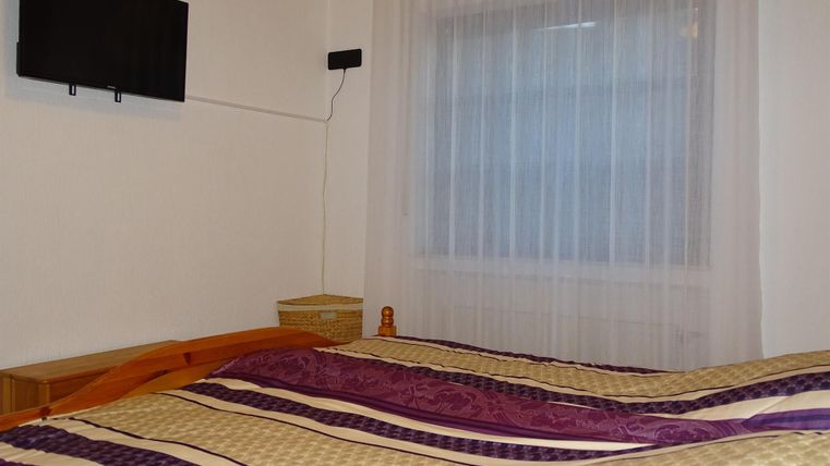 Ein gemütliches Schlafzimmer mit einem Doppelbett und bunten Bettlaken. An der Wand hängt ein Fernseher und die Fenster sind mit Vorhängen bedeckt.