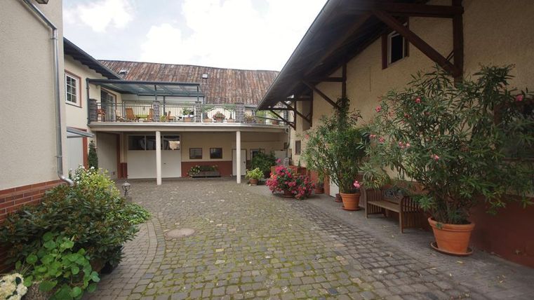 Ein gemütlicher Innenhof mit gepflastertem Boden und bepflanzten Töpfen. Im Hintergrund sind Gebäude mit einem glasüberdachten Bereich zu sehen.