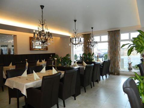 Ein elegantes Restaurant mit dunklen Holzmöbeln und stilvollen Kronleuchtern. Pflanzen und dekorative Vorhänge schaffen eine gemütliche Atmosphäre.