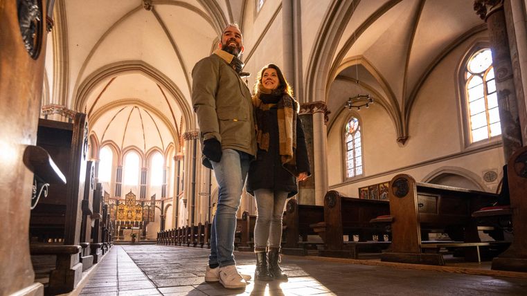 Un couple se tient dans une église historique et lumineuse. Les hauts plafonds et les belles fenêtres confèrent une atmosphère particulière à l'espace.