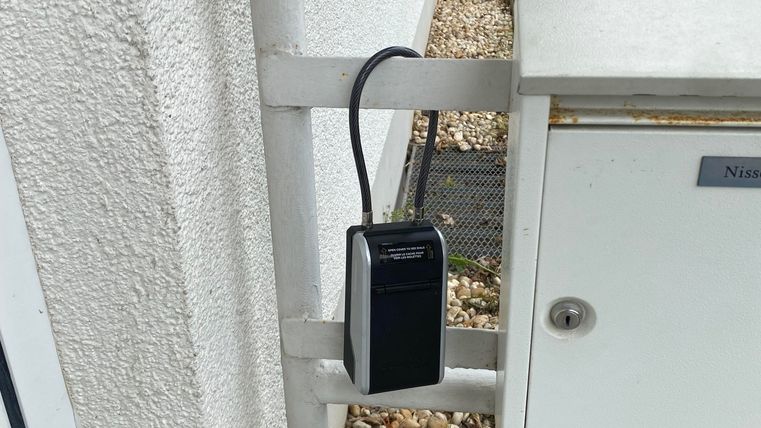 Ein schwarzes Schloss hängt an einem weißen Geländer neben einem Briefkasten. Der Hintergrund zeigt einen kleinen Kiesbereich und eine grüne Umgebung.