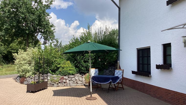 Eine gemütliche Terrasse mit einem grünen Sonnenschirm und einem Tisch. Im Hintergrund sind Pflanzen und ein weißes Haus sichtbar.