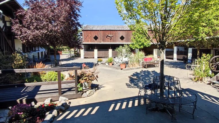 Ein malerischer Innenhof mit blühenden Pflanzen und Bäumen. In der Mitte befindet sich eine Sitzgelegenheit und im Hintergrund sind historische Gebäude sichtbar.