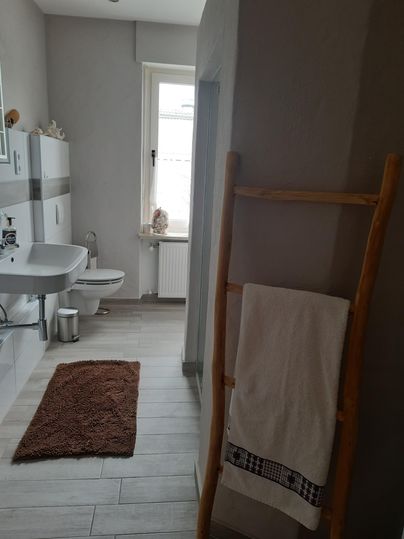 Ein modernes Badezimmer mit einem Waschbecken, einer Toilette und einem großen Fenster. An der Wand hängt ein Handtuchhalter aus Holz mit einem weißen Handtuch.