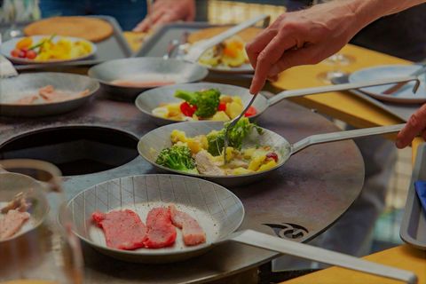 Zwei Hände bereiten frische Zutaten in kleinen Pfannen vor. Es gibt Fleisch, Gemüse und eine bunte Auswahl an Beilagen.