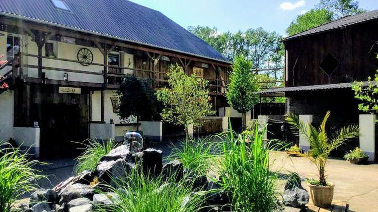 Ein Innenhof mit einem Wasserfall aus Steinen und üppigem Grün. Im Hintergrund sind rustikale Gebäude und Bäume zu sehen.
