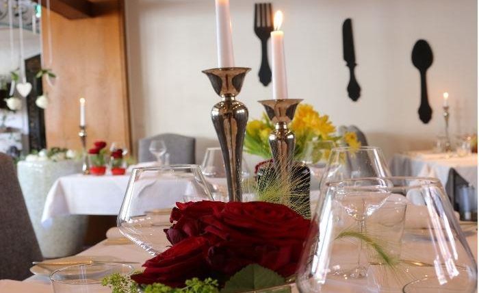 Ein elegant gedeckter Tisch mit Blumenarrangement und Kerzen. Im Hintergrund sind dekorative Essbestecke an der Wand zu sehen.