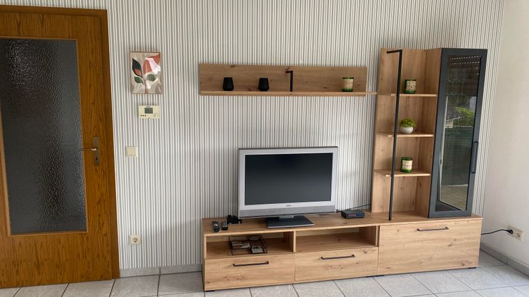 Ein Wohnzimmer mit einem modernen TV-Möbel aus Holz und einer Wandvitrine. Die Wände sind hell, und die Fliesen sind hellgrau.